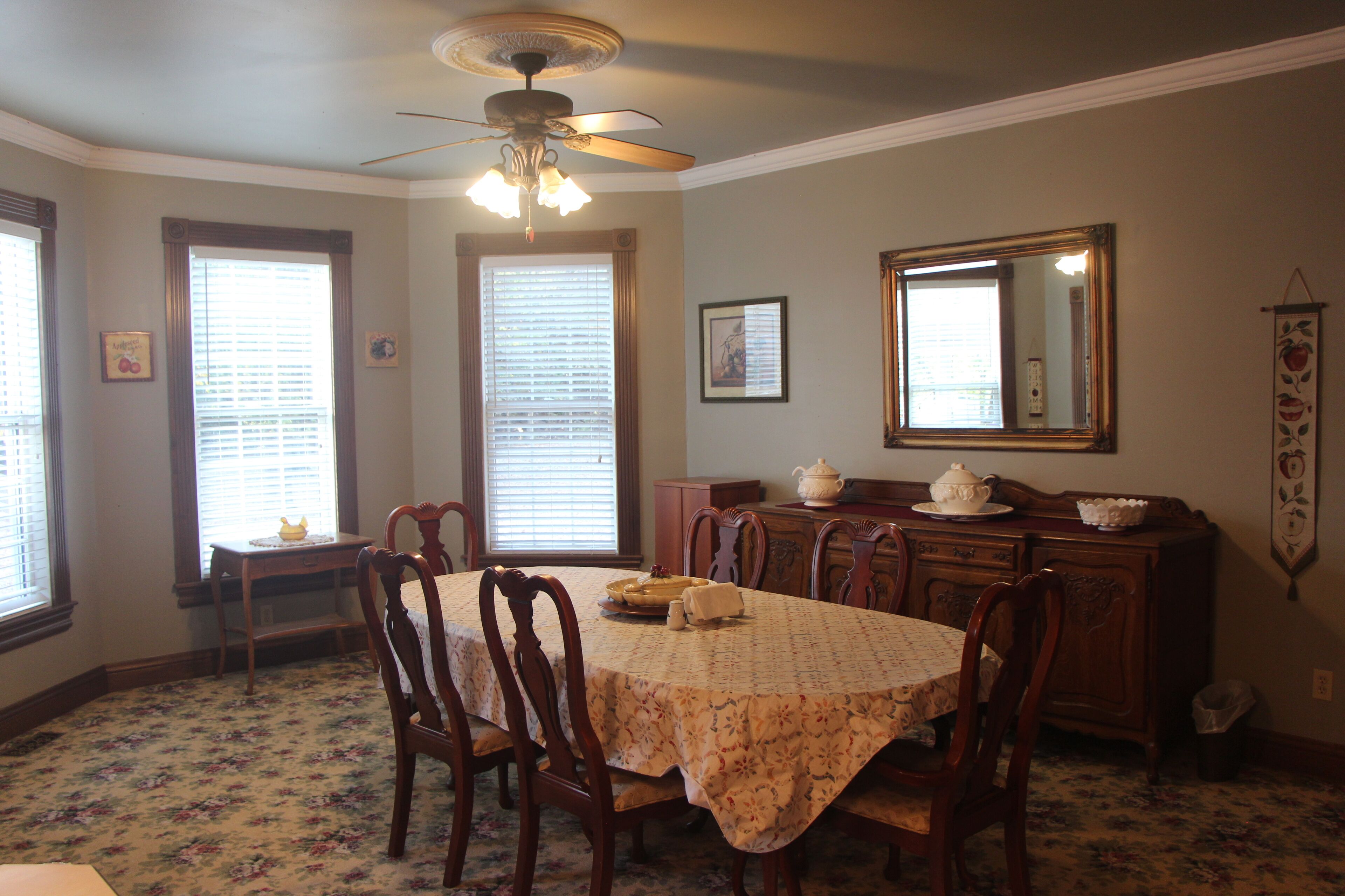 A dining room with a table set for four, surrounded by wooden chairs. A ceiling fan is visible above the table, and there is a large mirror on the wall.