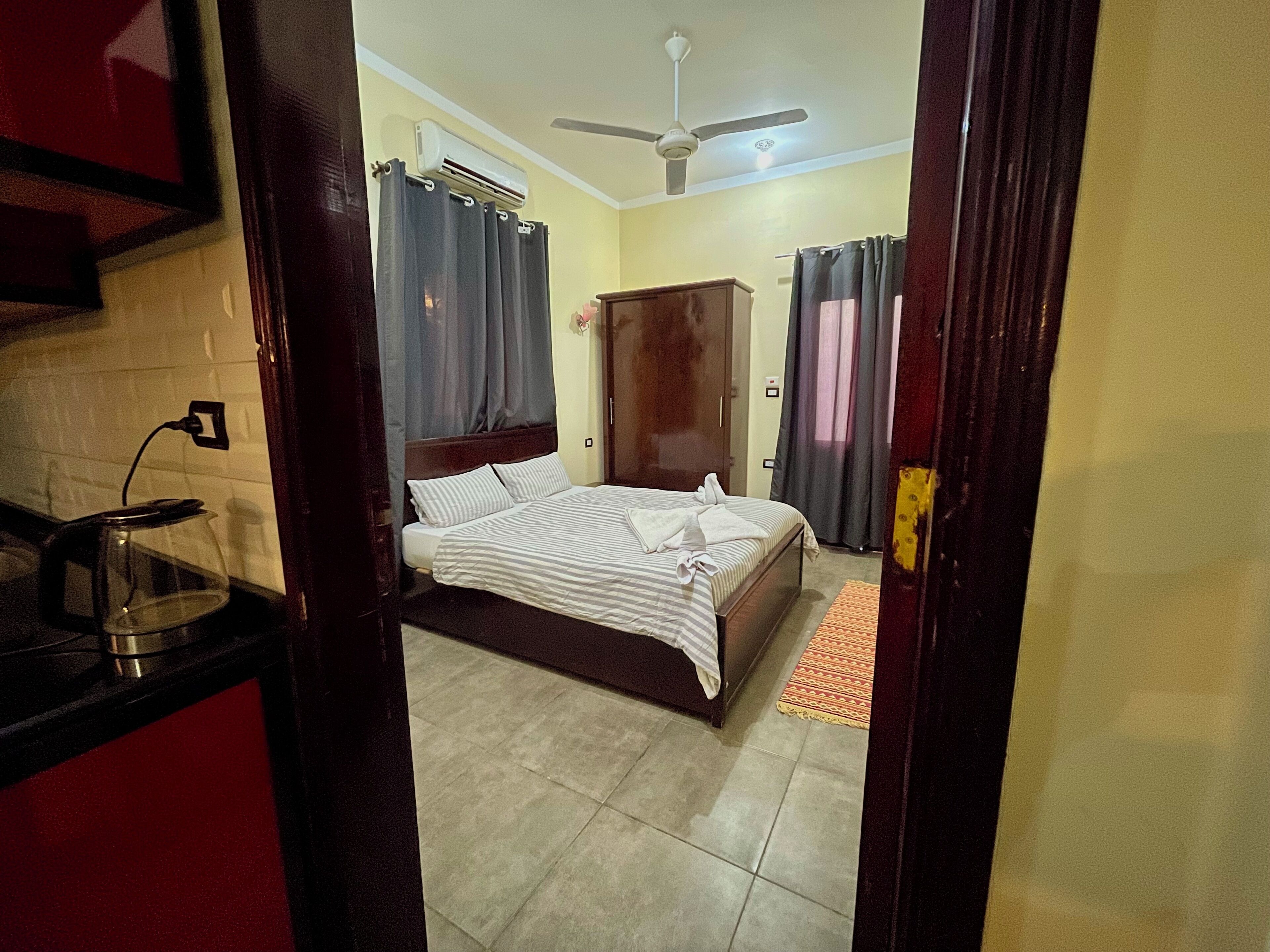 Habitación de hotel con cama, ventilador de techo, aire acondicionado y una pequeña cocina con hervidor.