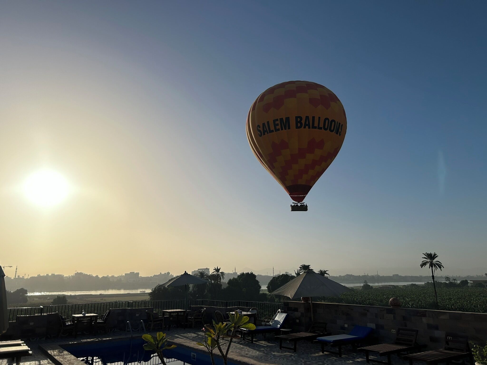A hot air balloon with "Salem Balloons" written on it, flying over a rooftop area with lounge chairs and a pool.