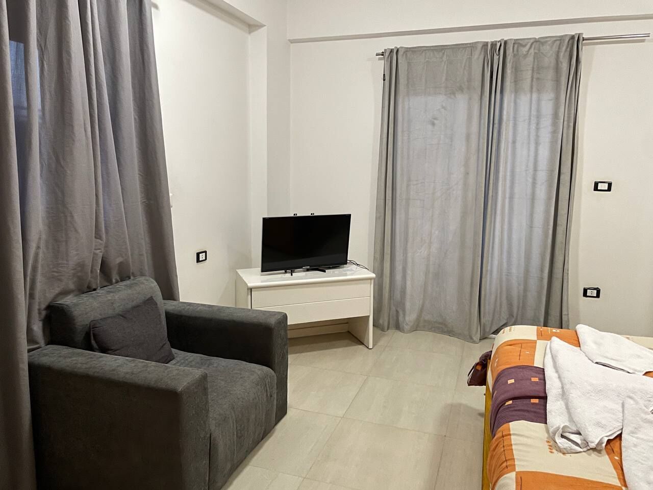 A bedroom with a grey sofa, a TV on a white table, a bed with a striped blanket, and grey curtains.