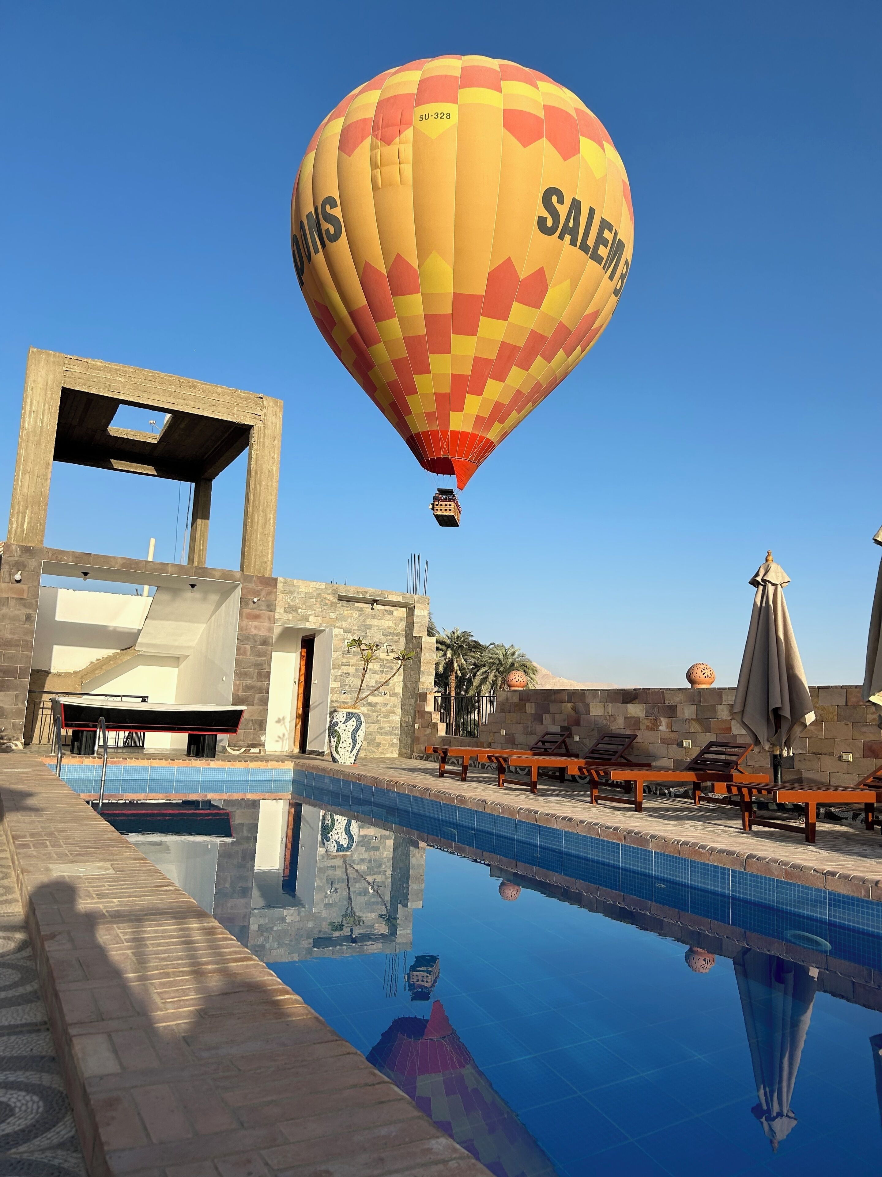 Une montgolfière sur laquelle on peut lire le mot « SALEM » survole une zone avec piscine, des chaises longues et un mur en pierre.