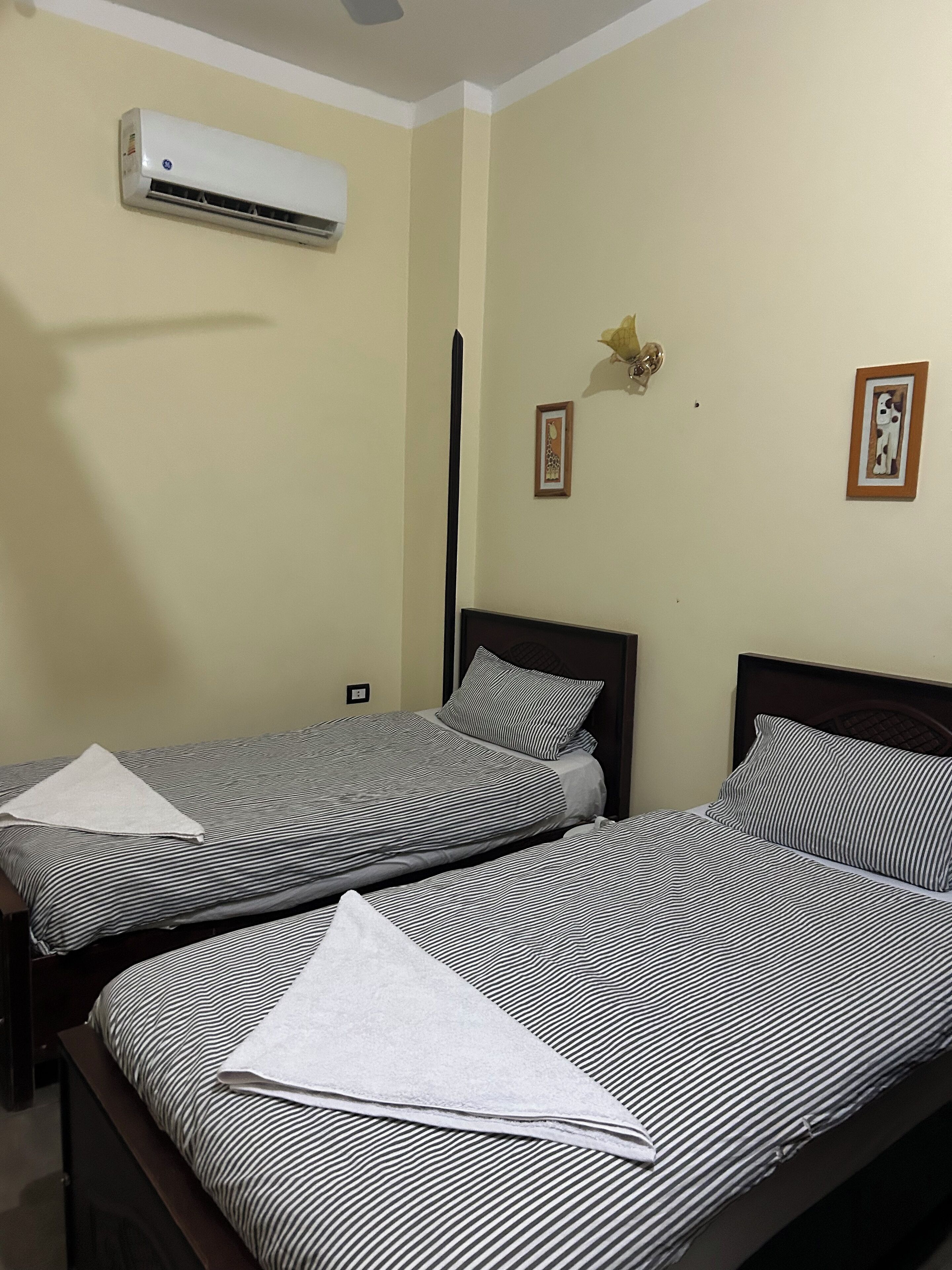 A hotel room with two beds, a ceiling fan, an air conditioning unit, and two framed pictures on the wall.