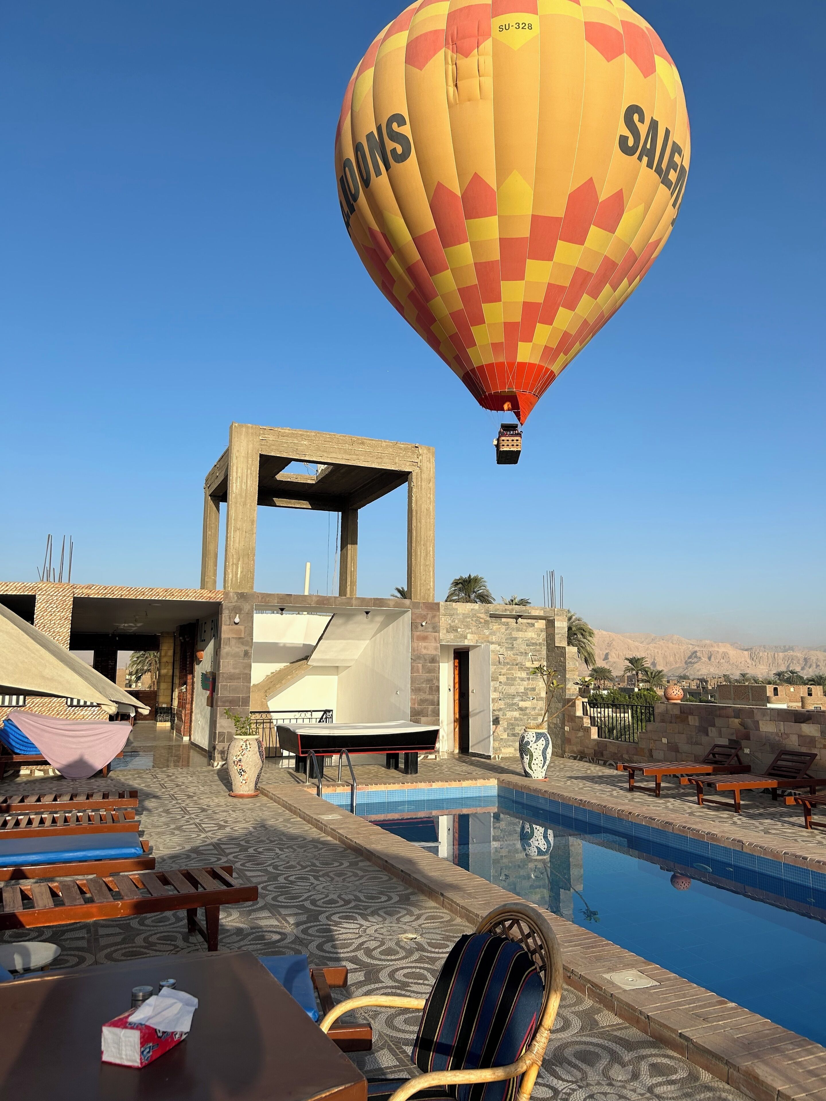 Une montgolfière sur laquelle est écrit le mot « SALON », une piscine, des chaises longues et, au loin, une vue sur le désert.