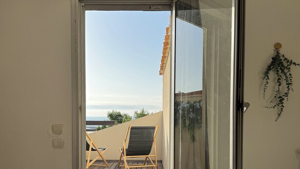 Un dormitori amb una porta corredissa de vidre que dona a un balcó de fusta, dues cadires i vista al mar.