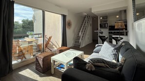 Un saló modern amb un sofà, una taula de centre i un otomà de vimetlla. Es veu una zona de menjador amb caires i una taula a través d'una porta de vidre. Una cuina amb una escala es troba al fons.
