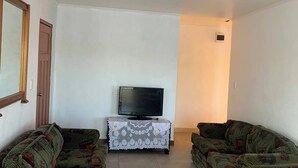 Ein Wohnzimmer mit zwei grünen Sofas mit Muster, einem Fernseher auf einem kleinen Tisch und einer Tür links.