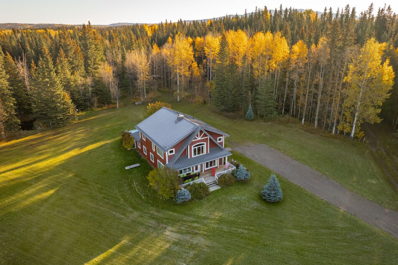 A two-story house with a red facade and a grey roof, surrounded by a well-maintained lawn and a dense forest.