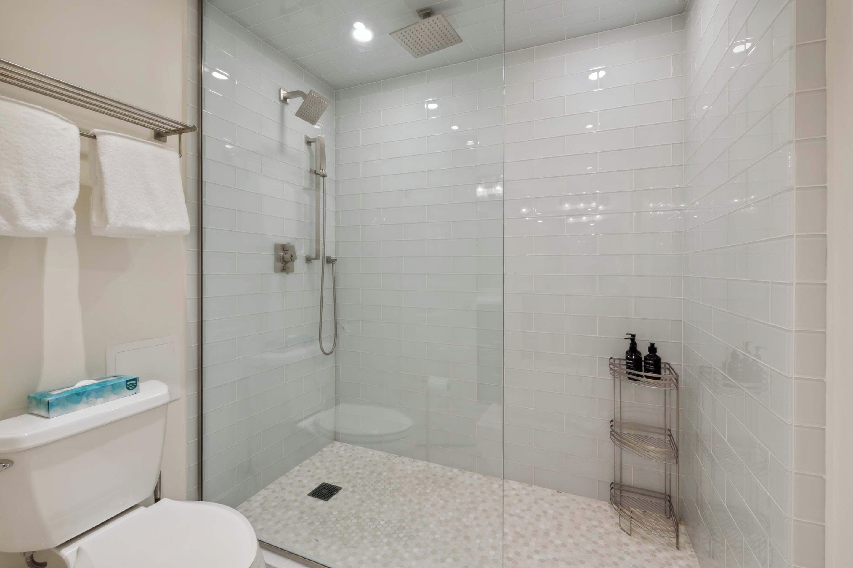 Un baño moderno con ducha de vidrio, paredes de azulejos blancos, un inodoro y un estante con artículos de aseo.