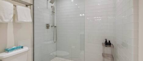 Un baño moderno con ducha de vidrio, paredes de azulejos blancos, un inodoro y un estante con artículos de aseo.