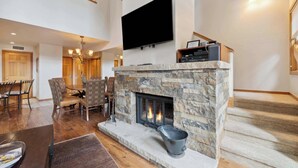 Amplia sala de estar con chimenea de piedra, un televisor de pantalla plana montado encima y una zona de comedor con sillas de mimbre.