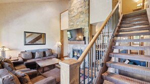 A spacious living room with a stone fireplace, a wooden staircase, and a large sofa with patterned cushions. - Woodrun Place 11 Three Bedroom Residence (Snowmass)