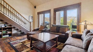 A spacious living room with a wooden staircase, large windows with a view, and a variety of seating options. - Woodrun Place 11 Three Bedroom Residence (Snowmass)
