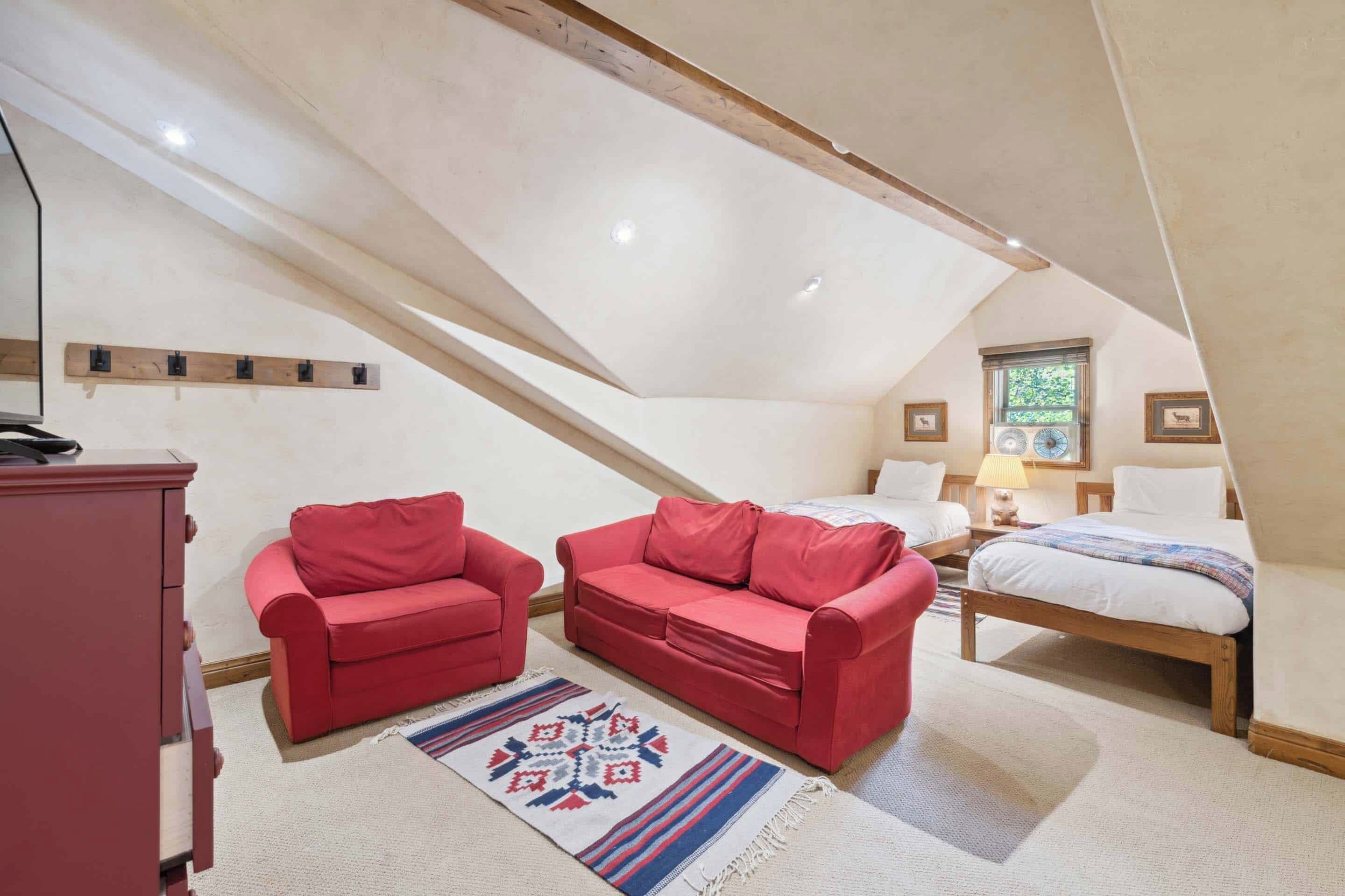 A cozy attic bedroom with a sloped ceiling, two red sofas, a bed, and a wooden dresser.
