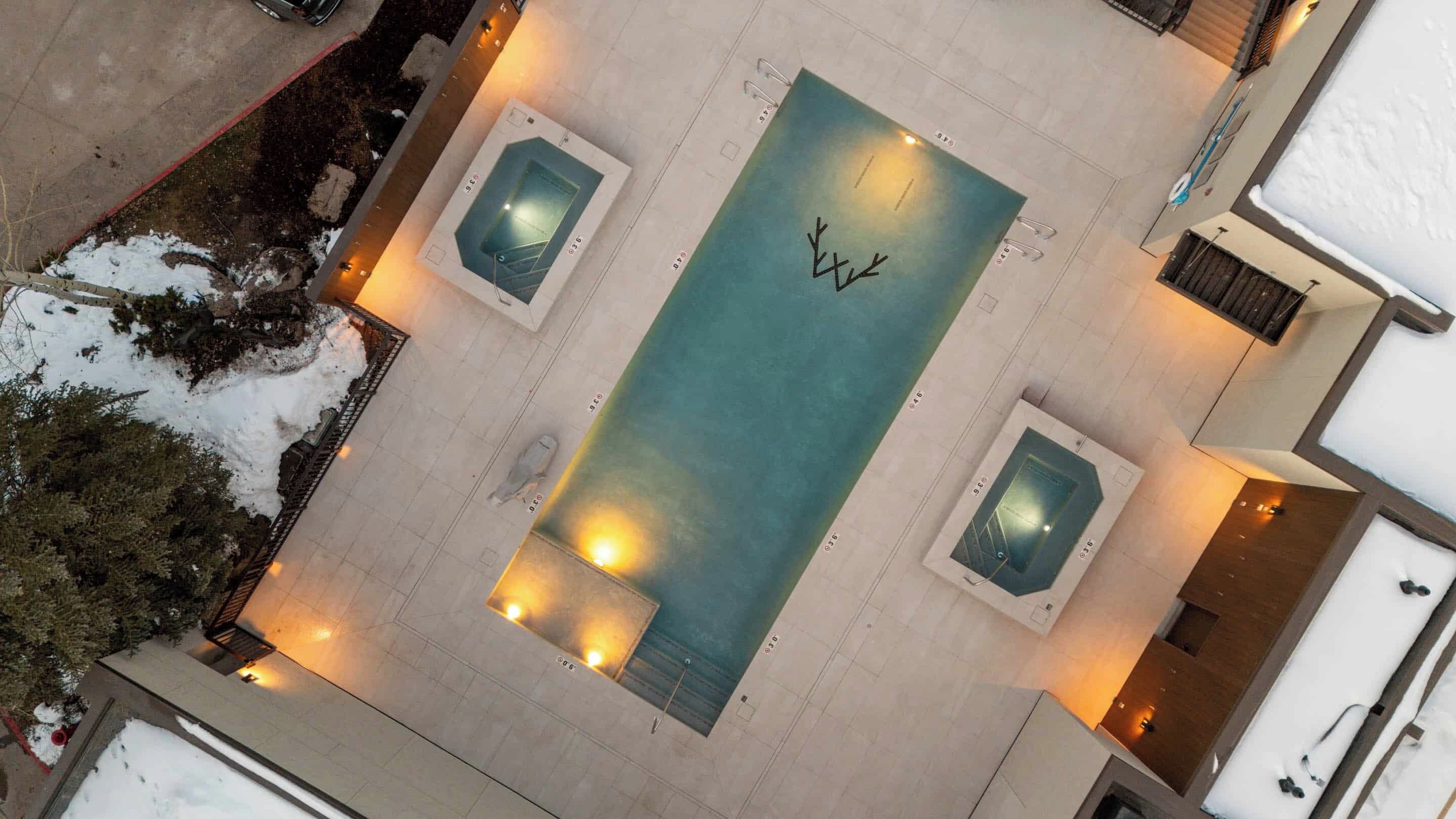 An aerial view of a hotel with a central swimming pool, two smaller pools, and a snow-covered landscape.