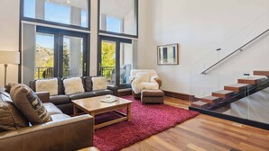 Uma sala de estar moderna com um sofá de couro, uma mesa de centro de madeira e um tapete vermelho. Há uma escada com guarda-corrimão de vidro e vista para o exterior.