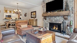 A living room with a stone fireplace, a television mounted on the wall, and a dining area in the background.
