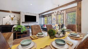A dining area with a wooden table set for six, surrounded by chairs. A fireplace with a stone facade is visible in the background, and a television is mounted on the wall. - Woodrun Place 20 Two Bedroom Residence (Snowmass)