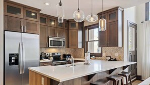 A modern kitchen with dark wooden cabinets, stainless steel appliances, a large island with stools, and pendant lighting. - Woodrun Place 32 Two Bedroom Residence (Snowmass)