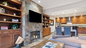 A modern living room with a fireplace, built-in shelves, a flat-screen TV, and a dining area with wooden cabinets and a kitchen in the background. - Woodrun Place 35 Two Bedroom Residence (Snowmass)