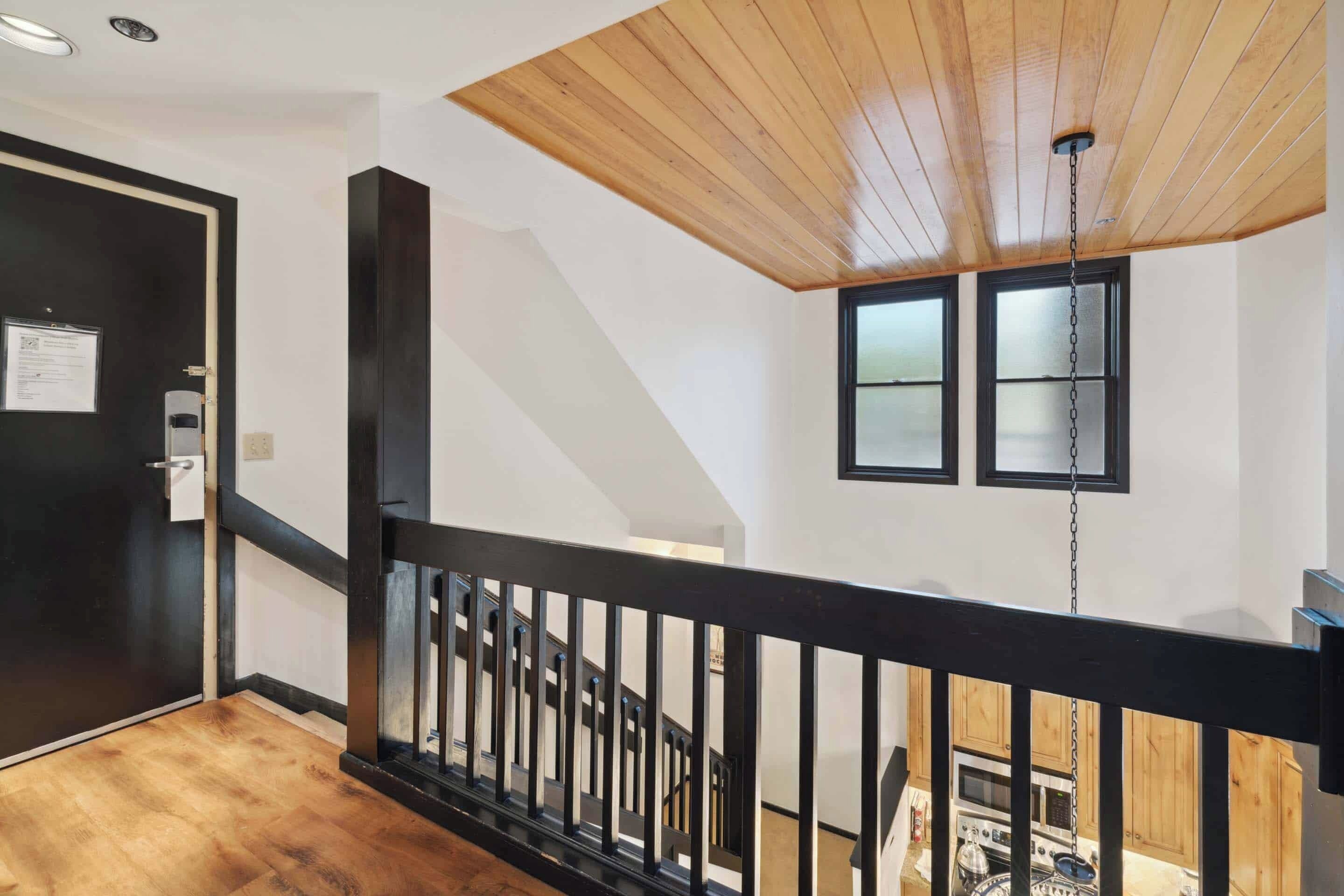 Ein modernes Interieur mit Holzboden, schwarzem Geländer und einer durch ein Fenster sichtbaren Küche.