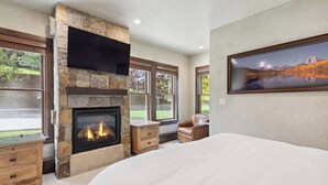 A bedroom with a stone fireplace, large windows, a bed, a nightstand, and a chair.