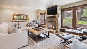 A spacious living room with a fireplace, a large TV mounted on a stone wall, and a view of the outdoors through large windows.