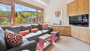A modern living room with a gray sofa, wooden cabinets, a TV, and a large window overlooking a scenic view.