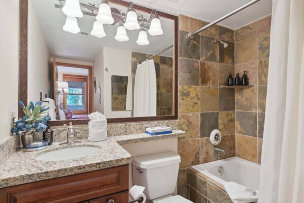Une salle de bain avec un lavabo surmonté d’un plan de travail en granit, des toilettes blanches, une baignoire et une douche aux carreaux en pierre.