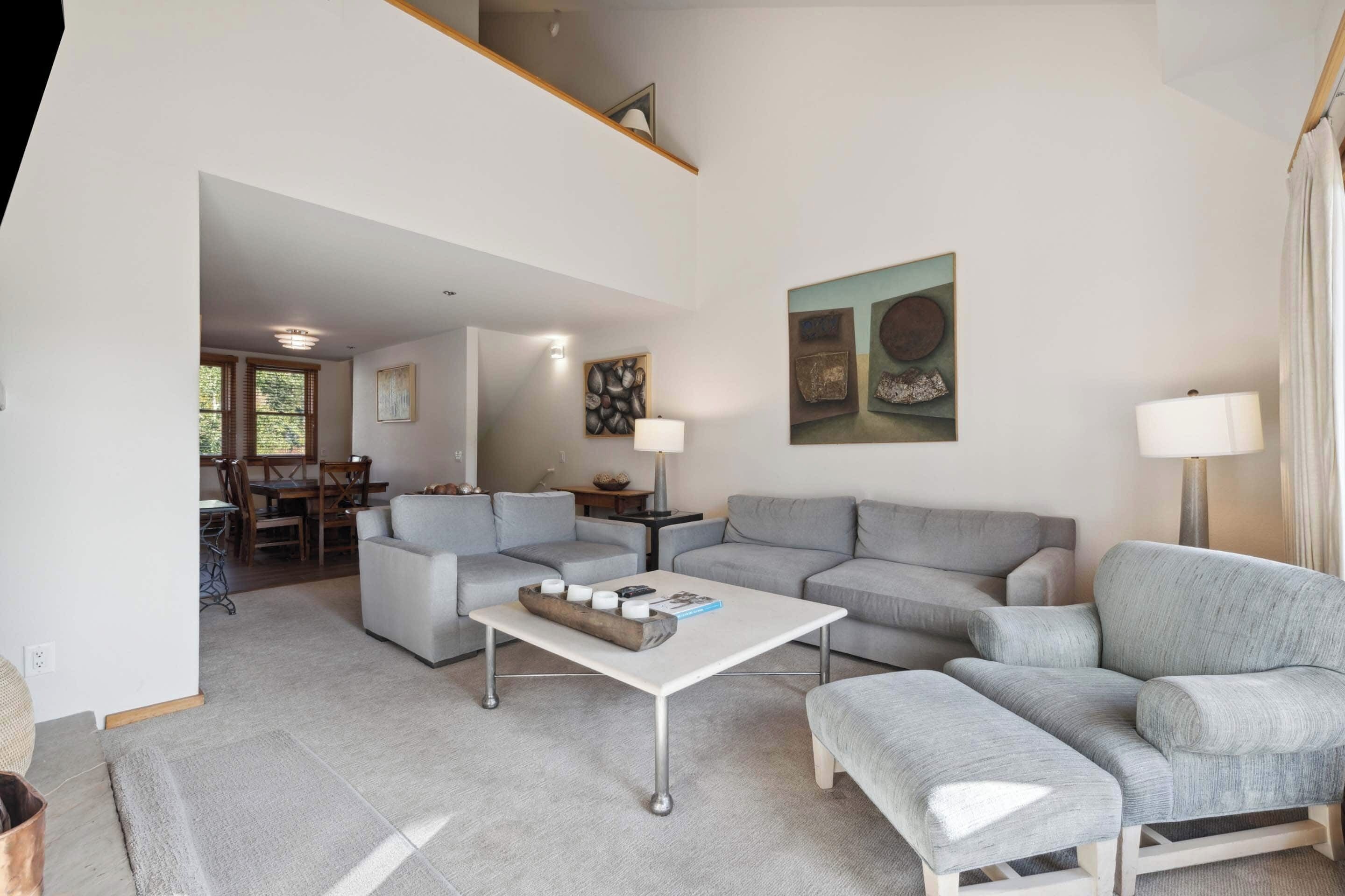 A spacious living room with a high ceiling, grey sofas, a white coffee table, and a dining area in the background.