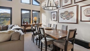 A dining area with a wooden table and chairs, framed artwork on the walls, and a view of the outdoors through large windows.