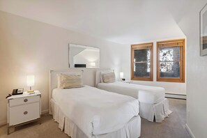 A bedroom with two beds, a nightstand with a lamp, a mirror, and two windows with wooden frames.