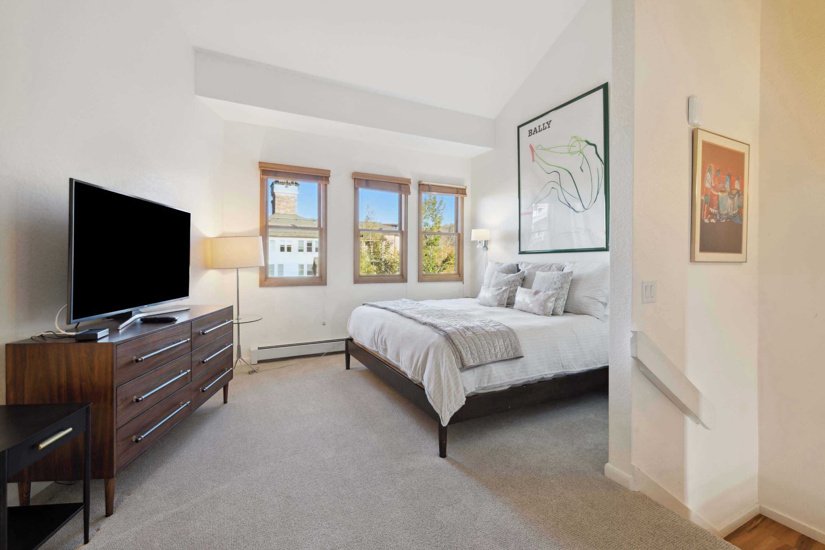 Un dormitorio con una cama, un televisor sobre un aparador de madera y una ventana con vistas a edificios.