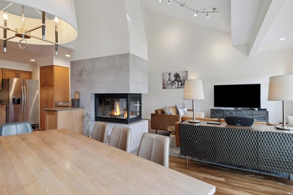 A modern living room with a fireplace, a dining area, and a kitchen with stainless steel appliances.