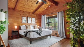 Quarto com teto de madeira, cama com roupa de cama branca, mesa de cabeceira com abajur, telescópio e uma grande janela com cortinas.