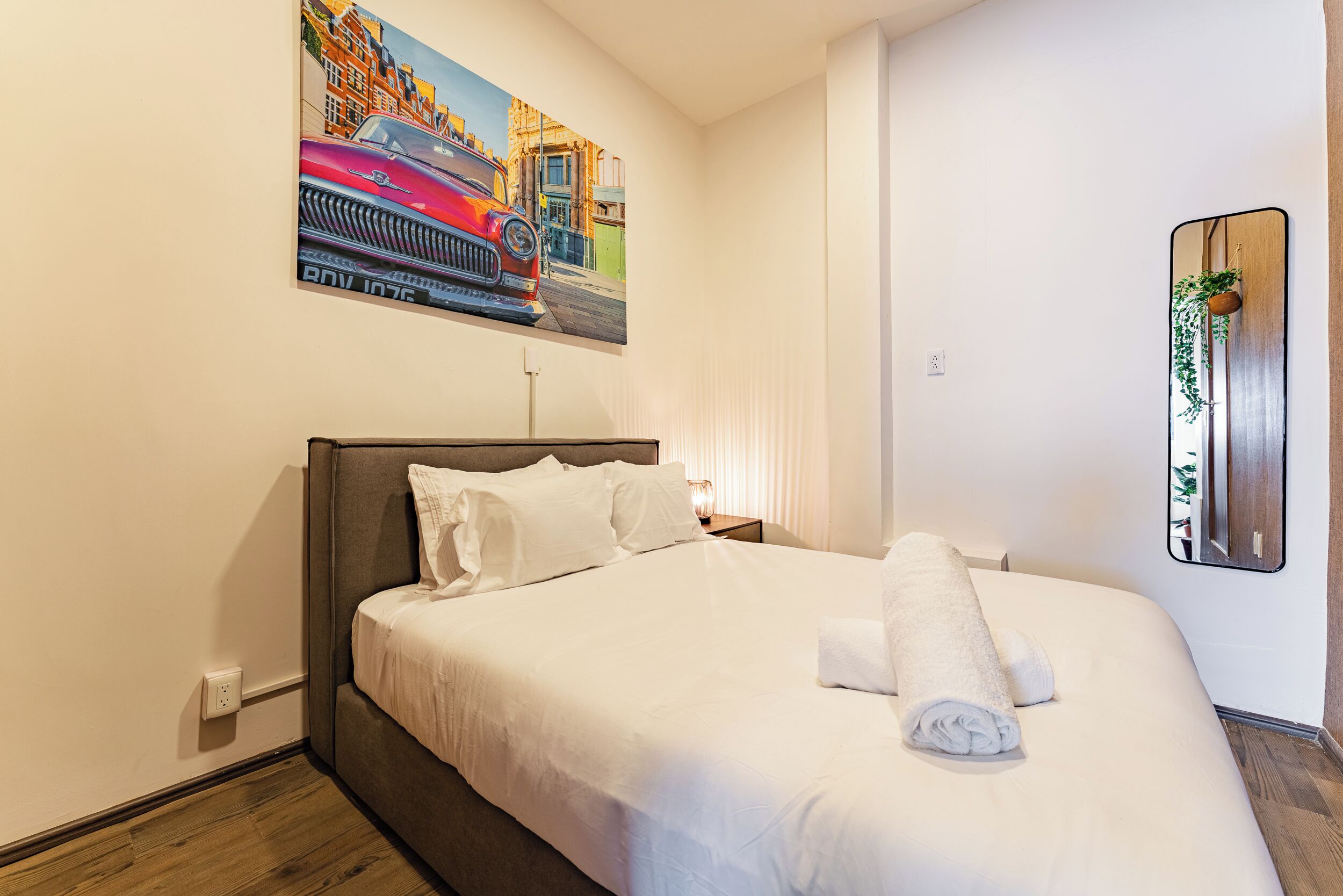 A neatly made bed with white linens and a rolled towel, a painting of a red car on the wall, and a mirror with a plant.