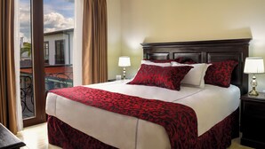 Habitación de hotel con una cama grande, dos mesitas de noche con lámparas, y un balcón que ofrece vistas a edificios y palmeras.