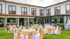Montaje para una boda al aire libre con mesas y sillas redondas, un área cubierta y un edificio con ventanas y balcones grandes.
