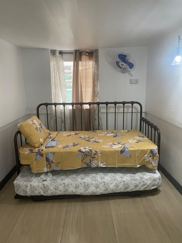 A single bed with a floral bedspread, a window with sheer curtains, and a wall-mounted fan.