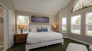 A bedroom with a large bed, two bedside tables with lamps, a wall-mounted TV, and three windows with blinds. - Pool, Pool Table, Board Games + Butterfly Garden (McKinney)