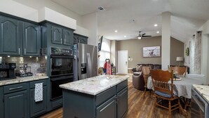 A modern kitchen with dark cabinets, a large island, and a dining area with wooden chairs and a table. - Pool, Pool Table, Board Games + Butterfly Garden (McKinney)