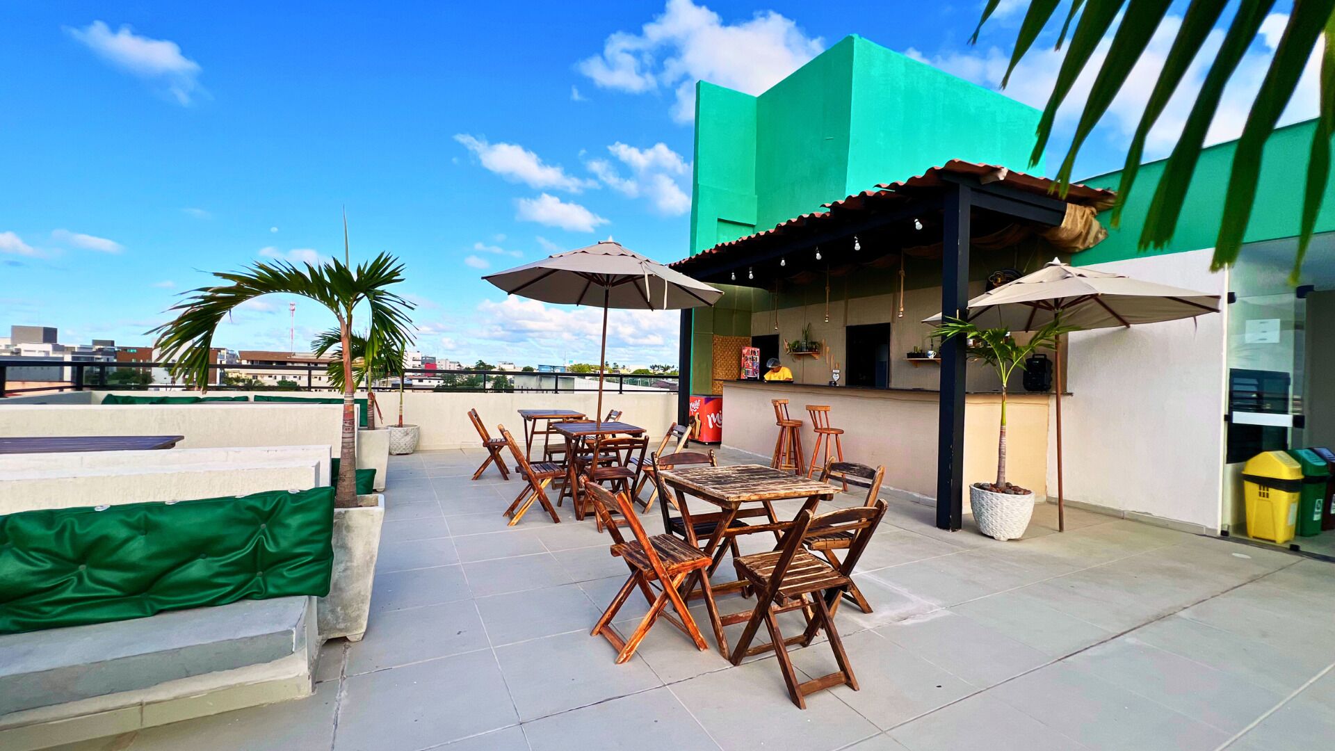 Un patio al aire libre con mesas y sillas de madera, sombrillas y un edificio verde al fondo.