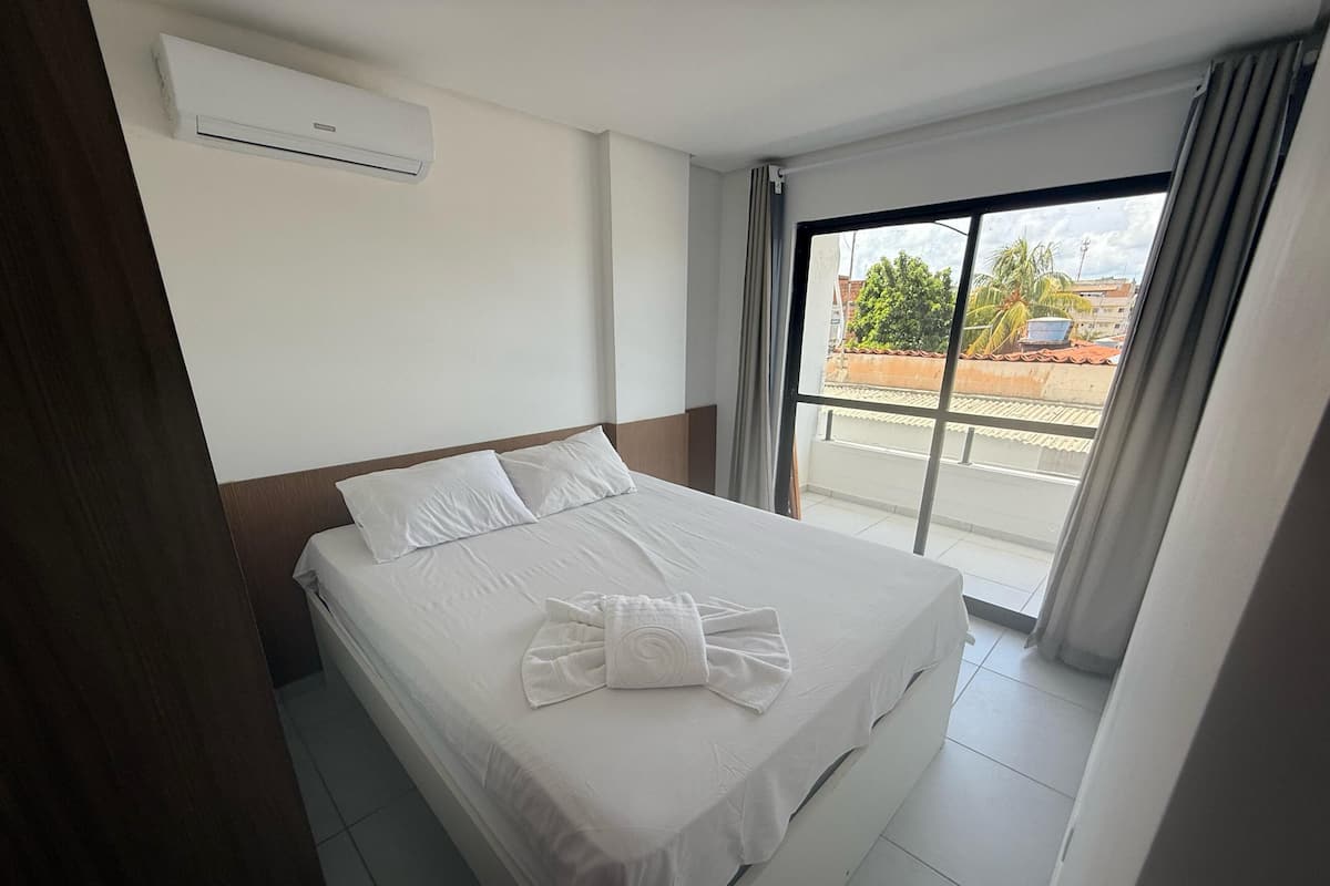 A hotel room with a bed, white linens, a window with curtains, and an air conditioning unit on the wall.