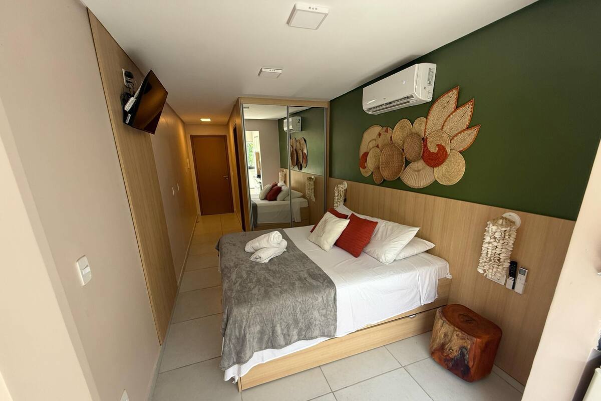 A hotel room with a bed, a TV mounted on the wall, an air conditioner, and a decorative wall hanging.