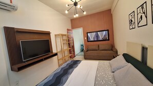 Et kompakt hotelværelse med et vægmonteret fladskærms-tv, en sofa, en seng med mønstret sengetøj og en loftlampe.