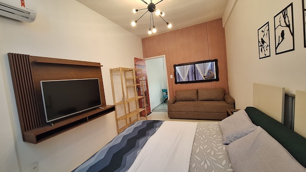 A compact hotel room with a flat-screen TV mounted on the wall, a sofa, a bed with patterned bedding, and a ceiling light fixture.