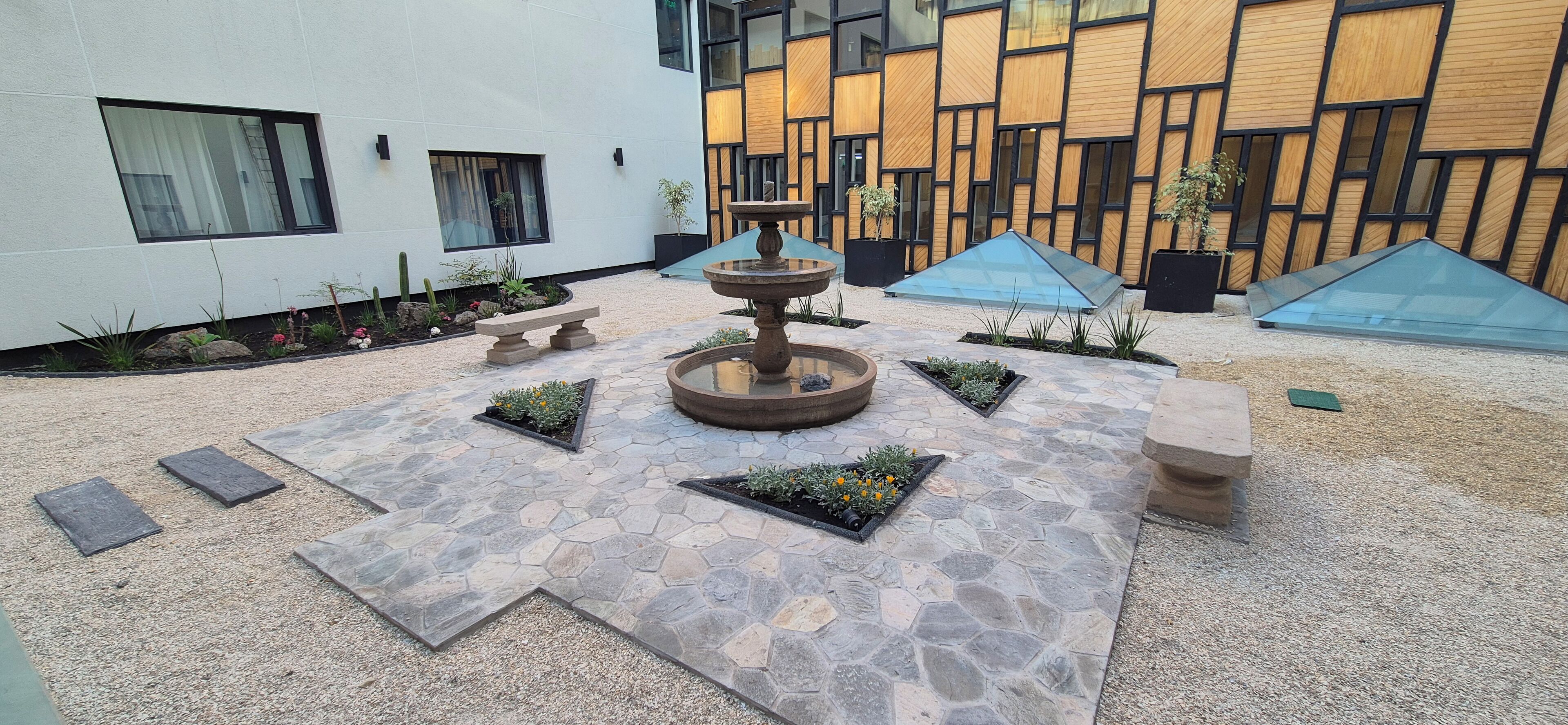 Une cour intérieure avec une fontaine centrale, des jardinières en pierre ornées de fleurs, un banc et, au fond, un bâtiment à l’architecture moderne.