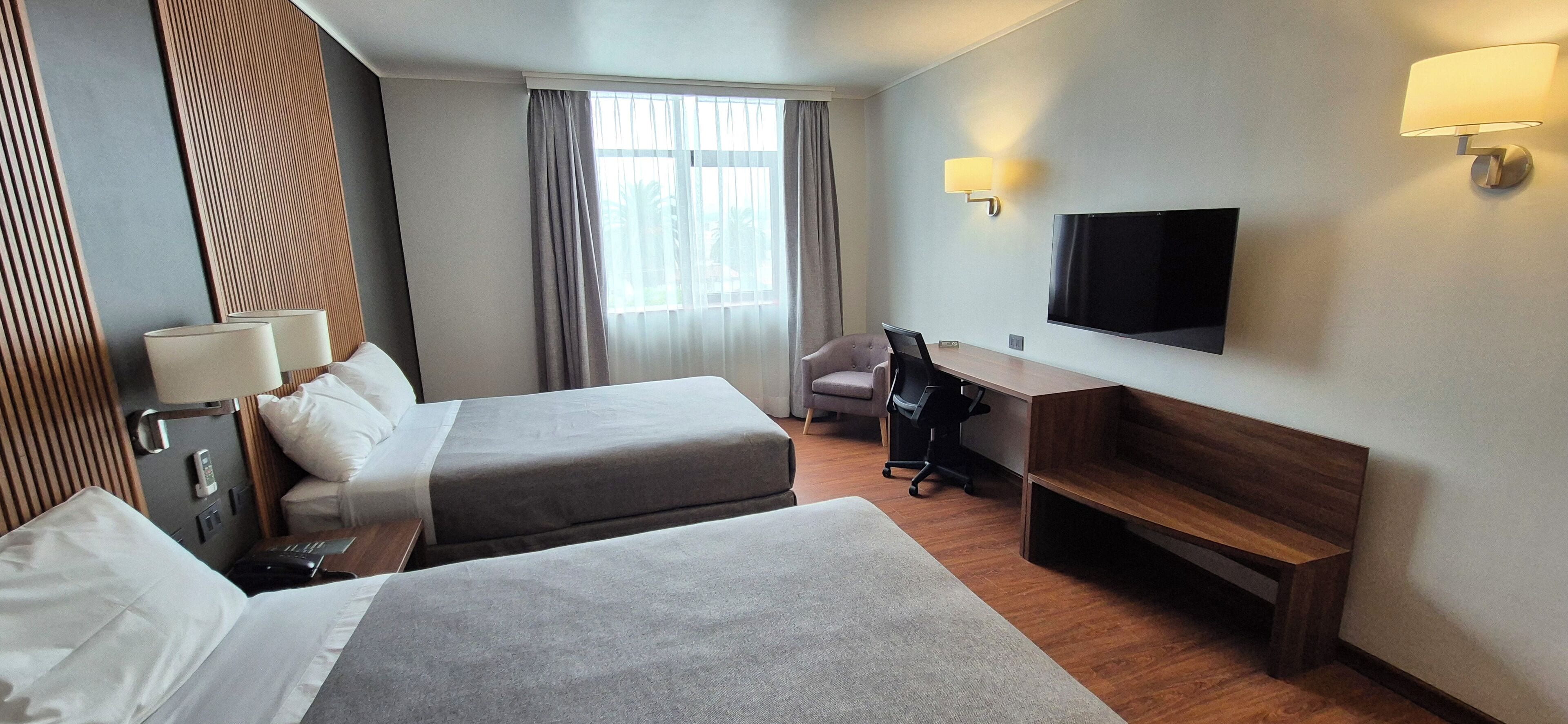 Une chambre d’hôtel avec deux lits, un bureau, une chaise, une télévision et une fenêtre avec des rideaux.