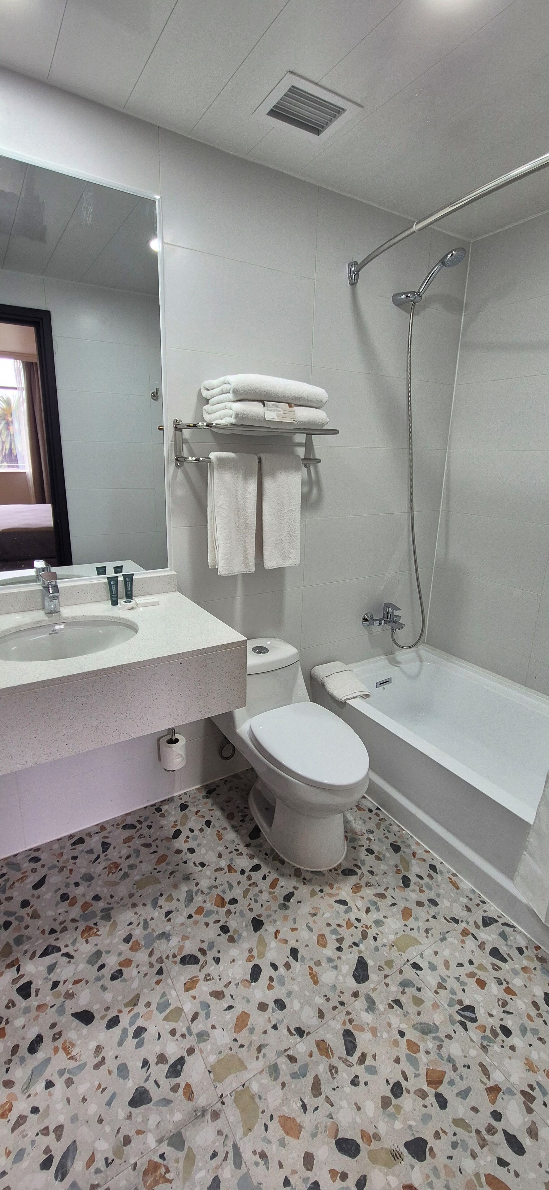 A bathroom with a white sink, toilet, and bathtub. There is a towel rack with folded towels above the toilet. The floor has a terrazzo pattern.