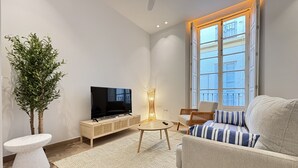 A modern living room with a flat-screen TV, a sofa with striped cushions, a small round table, a floor lamp, and a large window with a view of buildings. - Zarzuela (Málaga)