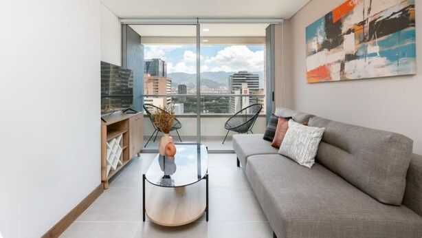 Una sala de estar moderna con un sofá, una mesa de centro de vidrio y vistas de la ciudad a través de amplias ventanas.